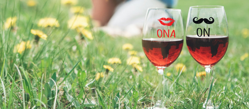 Slika čaša za vino na livadi, pikniku. Jedna čaša ima tisak brka i napisano on dok druga čaša ima otisnute usne te ispod riječi ona